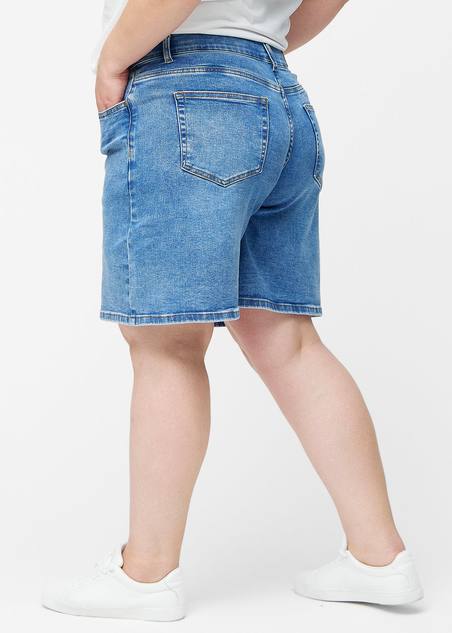 Løse, klassisk blå denim shorts i farven Rivers set fra siden på plus-size model, hvor den brede pasform og længden til lige over knæet er synlig.