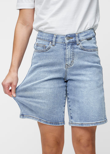Model dehnt die lockeren, hellblauen Denim-Shorts in der Farbe Waves an der Hüfte, um den Stretch des Materials zu zeigen.