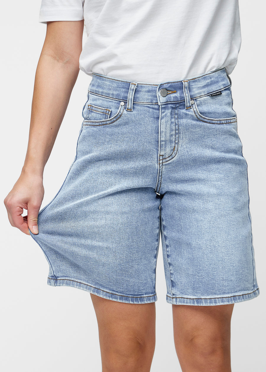 Model dehnt die lockeren, hellblauen Denim-Shorts in der Farbe Waves an der Hüfte, um den Stretch des Materials zu zeigen.
