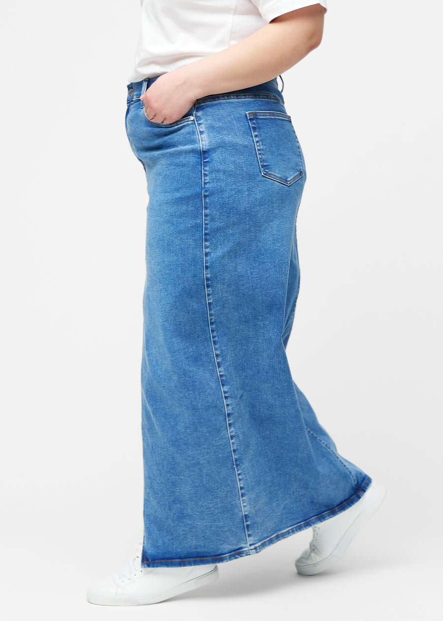 Lang, klassisk blå denimnederdel i farven Rivers med regulær pasform set fra siden på plus-size model, med synligt slids foran.