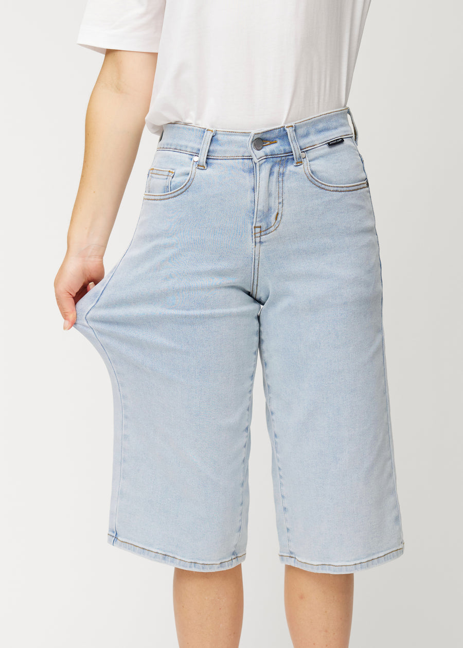 Ein Model dehnt die lockeren, hellblauen Waves Denim-Capris an der Hüfte, um den Stretch des Materials zu zeigen.