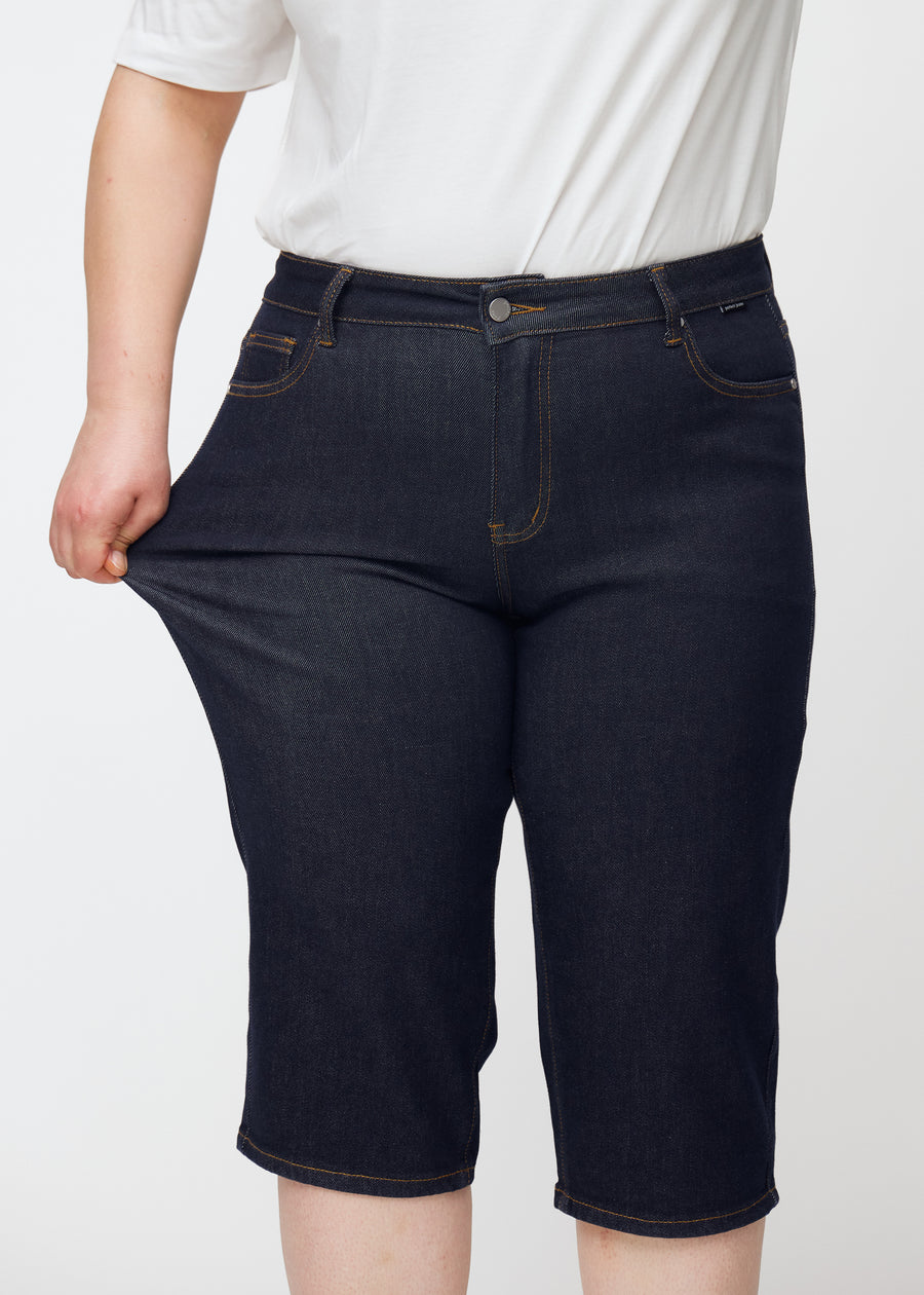 Plus-Size-Model dehnt tief dunkelblaue Denim-Capris für Damen in der Farbe Midnights am Oberschenkel, um den Stretch zu zeigen.