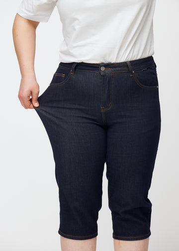 Plus-Size-Model dehnt tief dunkelblaue Denim-Capris für Damen in der Farbe Midnights am Oberschenkel, um den Stretch zu zeigen.