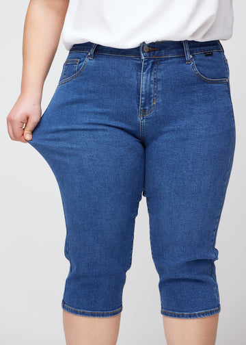 Slim-Capris für Damen aus dunkelblauem Denim, Farbe Oceans. Ein Plus-Size-Model dehnt den Stoff am Oberschenkel, um die Flexibilität zu zeigen.