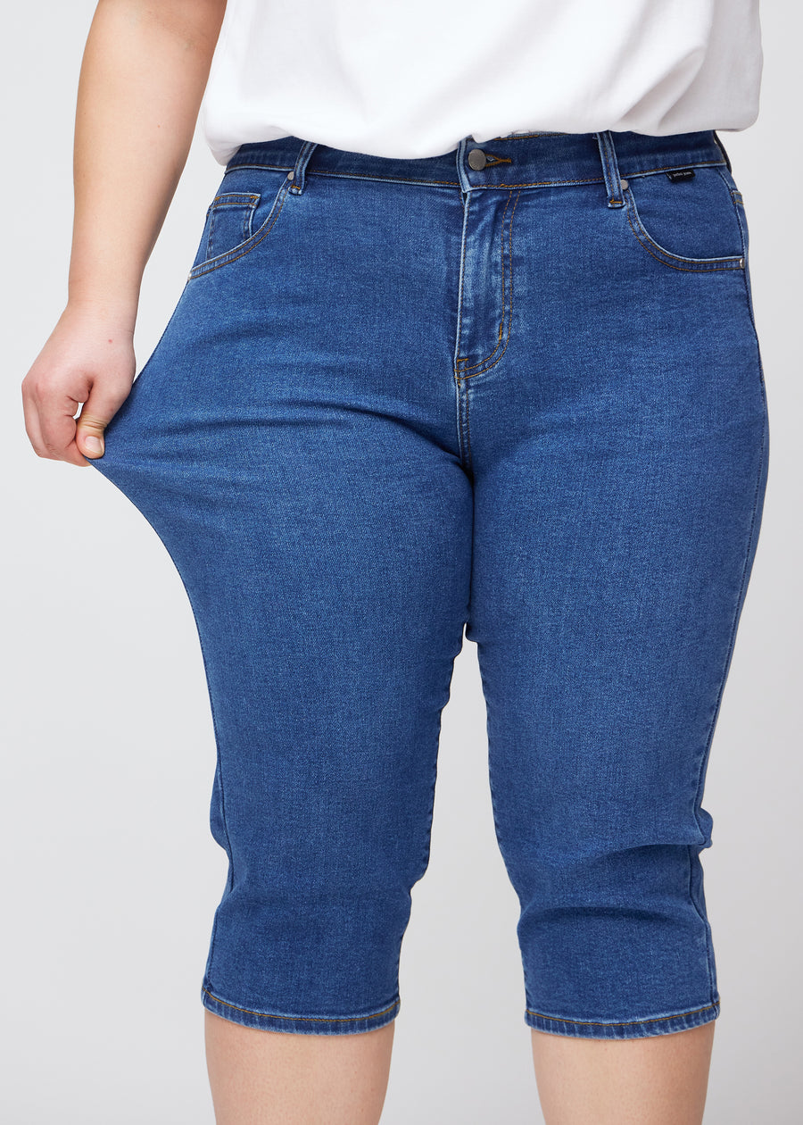 Slim-Capris für Damen aus dunkelblauem Denim, Farbe Oceans. Ein Plus-Size-Model dehnt den Stoff am Oberschenkel, um die Flexibilität zu zeigen.