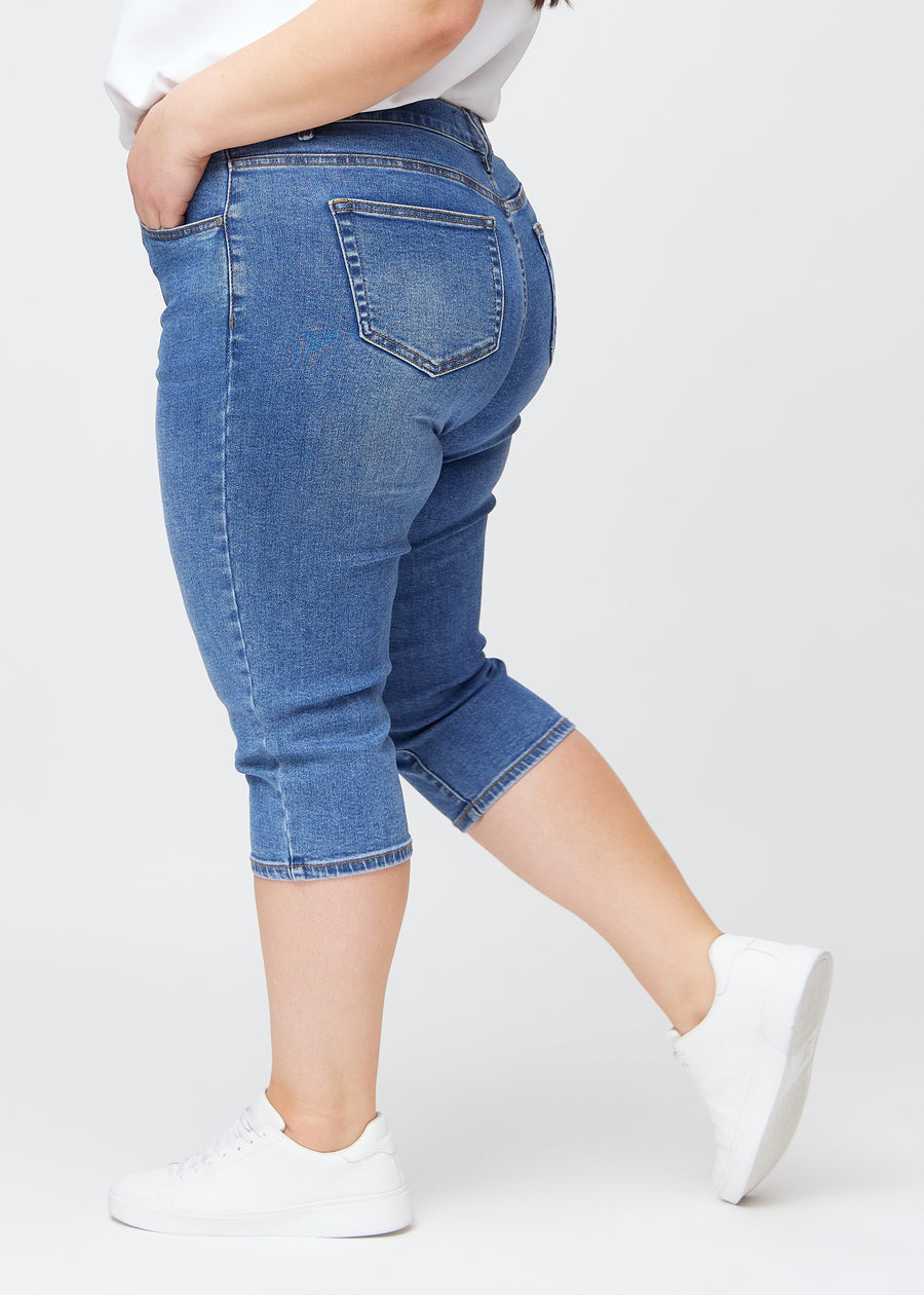Slim-Capris für Damen aus blauem Denim, Farbe Rivers. Seitenansicht an einem Plus-Size-Model, figurbetonter Schnitt und mittelhoher Bund (Mid Rise).