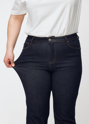 Ein Plus-Size-Model dehnt die tief dunkelblaue Denim jeans für Damen in der Farbe Midnights am Oberschenkel, um die Dehnbarkeit des Materials zu zeigen.