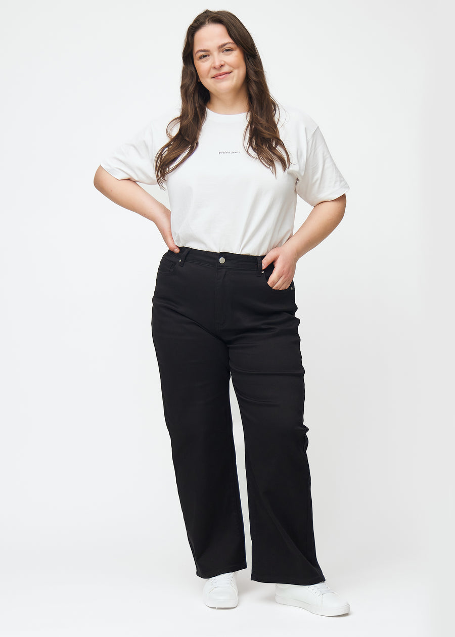 Ein Ganzkörperbild eines Plus-Size-Models in schwarzen hochtaillierten jeans, die an der Taille eng anliegen und eine lockere Passform mit Weite nach unten haben.