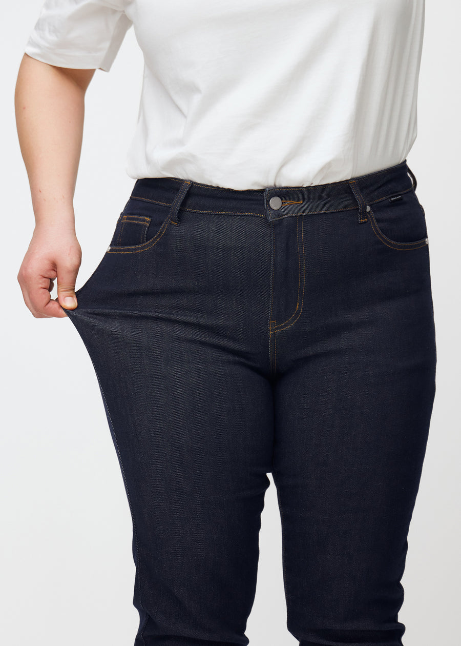 Ein Plus-Size-Model dehnt die tief dunkelblaue Denim jeans für Damen in der Farbe Midnights am Oberschenkel, um die Dehnbarkeit des Materials zu zeigen.
