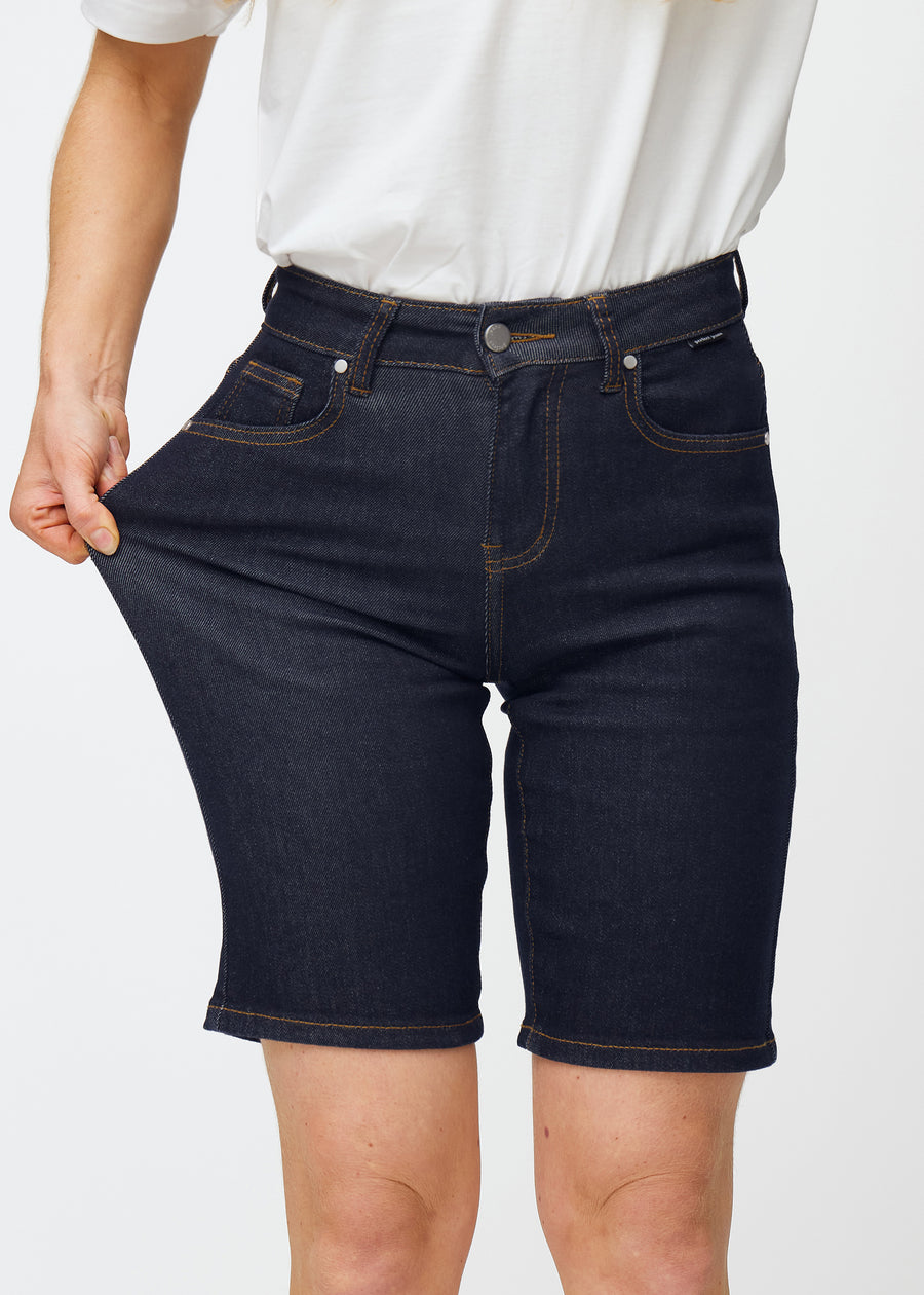 Model dehnt tief dunkelblaue Denim-Shorts für Damen in der Farbe Midnights am Oberschenkel, um den Stretch des Materials zu zeigen.