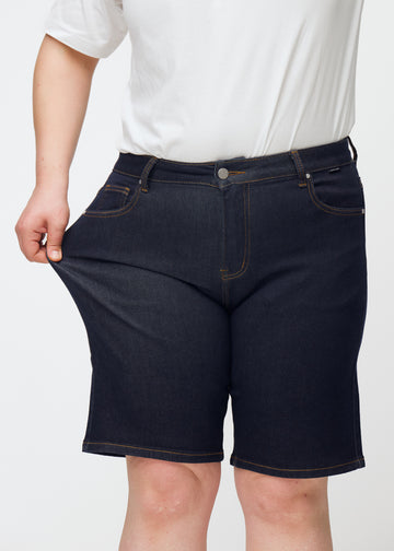 Plus-Size-Model dehnt tief dunkelblaue Denim-Shorts für Damen in der Farbe Midnights am Oberschenkel, um den Stretch zu zeigen.