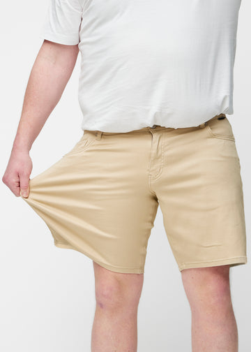 Plus-Size-Modell dehnt beige Shorts für Herren in der Farbe Gazelles am Oberschenkel, um den Stretch des Materials zu zeigen.