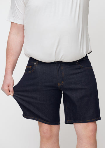 plus-size-model dehnt dunkelblaue denim shorts für herren in der farbe midnights am oberschenkel, um den stretch zu zeigen.